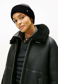 Jeune femme portant une veste en shearling noire, un pull rayé gris foncé, une chemise grise à boutons, un bandeau en tricot noir et de petites boucles d'oreilles en anneau.