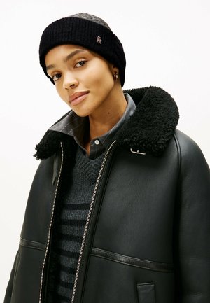 Jeune femme portant une veste en shearling noire, un pull rayé gris foncé, une chemise grise à boutons, un bandeau en tricot noir et de petites boucles d'oreilles en anneau.