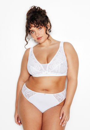 Donna dai capelli ricci che indossa un reggiseno di pizzo bianco e biancheria intima coordinata, in piedi davanti a uno sfondo bianco, che guarda verso la fotocamera.