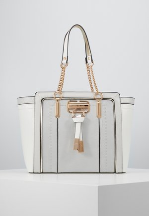 Sac cabas blanc et gris avec anses en chaîne dorée et garnitures décoratives dorées à l’avant, présenté sur un piédestal blanc sur fond gris.