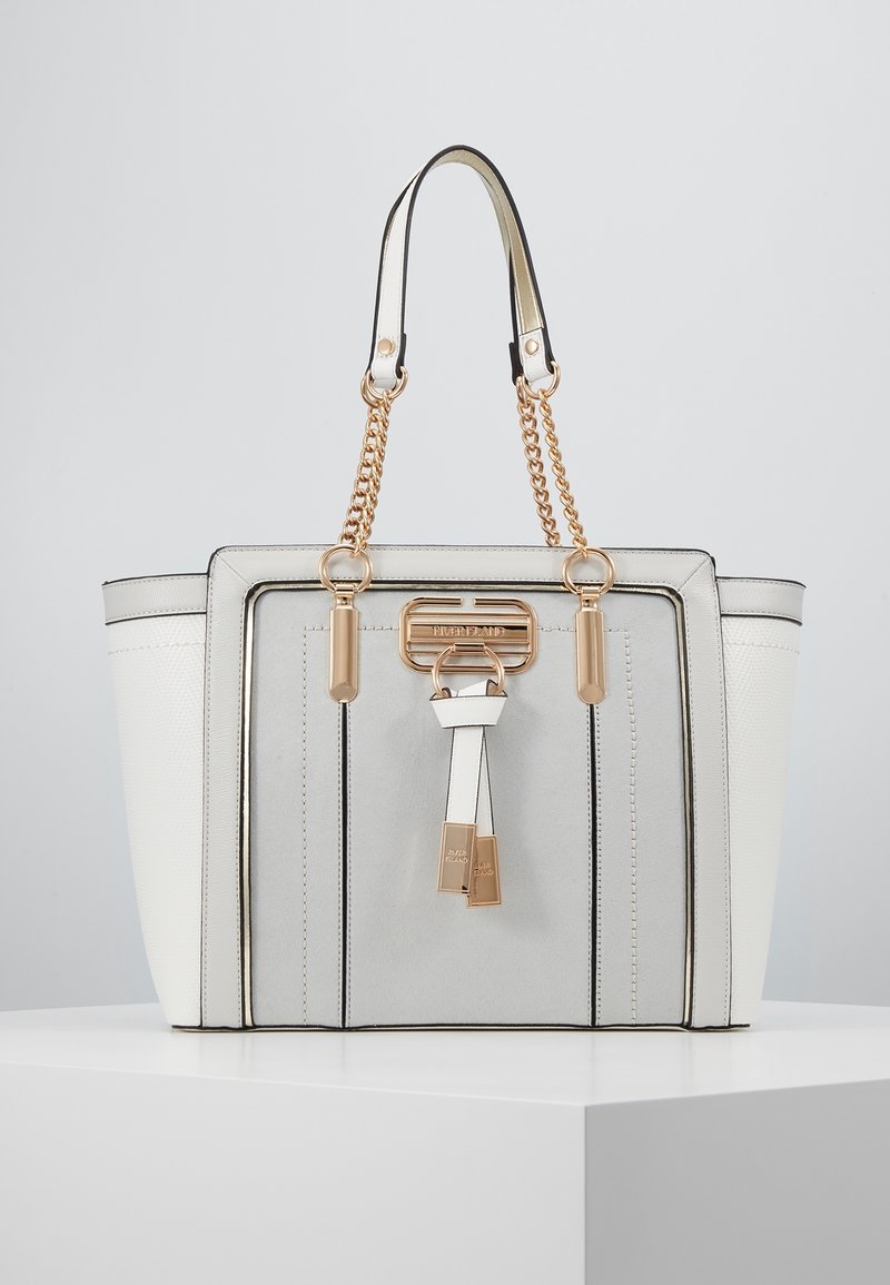 Sac cabas blanc et gris avec anses en chaîne dorée et garnitures décoratives dorées à l’avant, présenté sur un piédestal blanc sur fond gris.