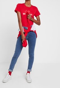 Camiseta roja de algodón con mangas cortas, combinada con jeans de mezclilla azules. Zapatillas deportivas blancas y calcetines rojos completan el look, con una camisa roja atada a la cintura.