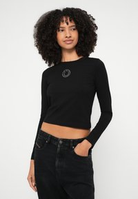 Čierne orezané tričko s dlhým rukávom a okrúhlym výstrihom, s bielym logom smajlíka v strede. Nosené s čiernymi džínsami s vysokým pásom.