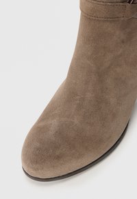 Bottine en suède taupe avec un bout arrondi et une texture lisse. Elle dispose d'un petit talon et d'une fine sangle autour de la cheville.