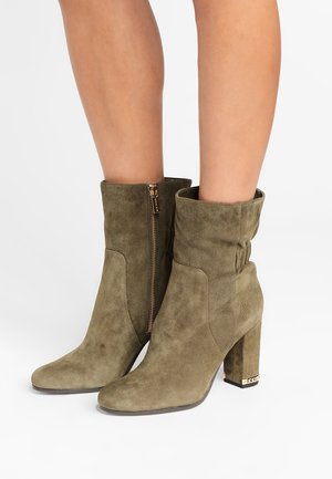 Femme portant des bottines en daim vert olive à talons blocs et fermetures éclair latérales, montrée de mi-mollet vers le bas sur fond blanc.