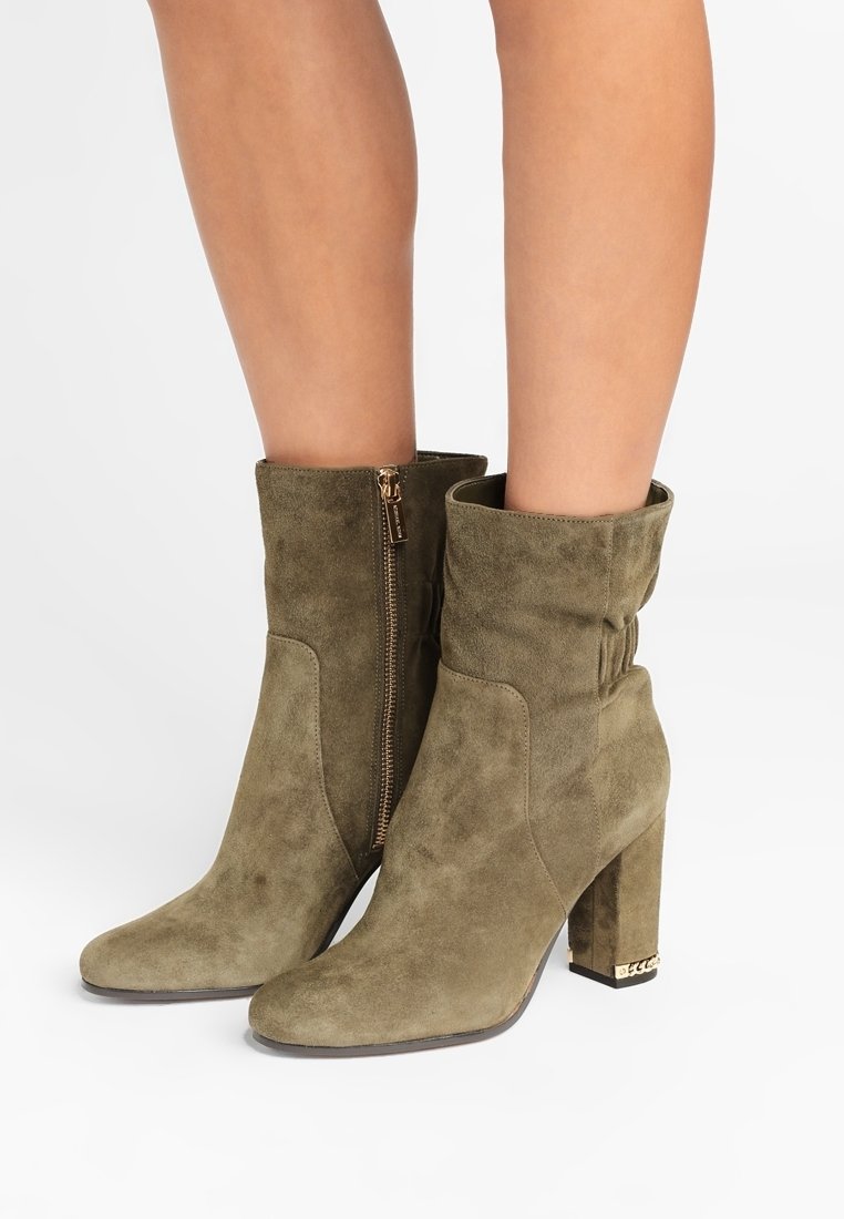 Femme portant des bottines en daim vert olive à talons blocs et fermetures éclair latérales, montrée de mi-mollet vers le bas sur fond blanc.