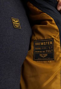 Granatowa teksturowana kurtka z żółtą satynową podszewką. Posiada czarną metkę z białym i złotym napisem "Brewster" oraz szczegóły produktu.