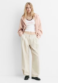 Helles rosa Zip-Up Hoodie, weißes, verkürztes Tanktop mit bedrucktem Text, beige weite Hose und schwarze Schuhe. Glatte Texturen und lockere Passform.