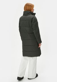 Lange, donkergroene gewatteerde jas met een hoge kraag, met horizontale naden en lange mouwen. Draagt over witte broek en zwarte schoenen.