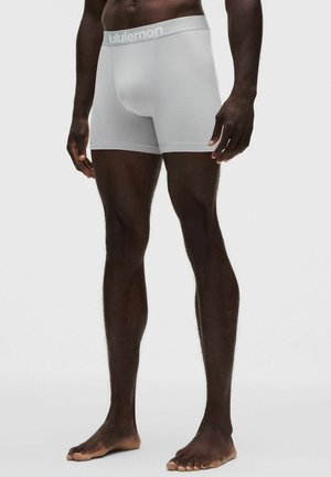 Persona de piel oscura que lleva puestos unos calzoncillos bóxer blancos de Lululemon, de pie descalza sobre un fondo claro y liso.