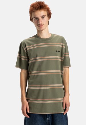 Jonge man met rood haar in een groen gestreept shirt en blauwe jeans, staande tegen een effen lichte achtergrond.