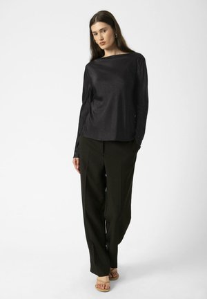 Žena s dlhými tmavými vlasmi, oblečená v čiernom tričku s dlhým rukávom, voľné čierne nohavice a béžové sandále s odhalenými prstami, stojaca na bielom pozadí.