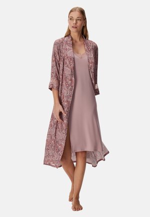 Vrouw staat blootsvoets en draagt een lichtroze nachthemd met kanten rand en een lange, patroonachtige kamerjas in tinten van roze en mauve.