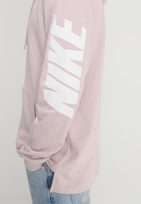 Sudadera Nike rosa con gran logo blanco en la manga, tela suave, bolsillo canguro, mangas largas y corte relajado. Diseño clásico de ropa deportiva.