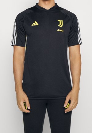 Person trägt ein schwarzes Juventus-Sporttrikot mit gelbem Adidas-Logo, Jeep-Sponsor und weißen Streifen an den Ärmeln, kombiniert mit schwarzen Hosen.