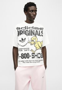 Νέος άνδρας που φοράει λευκό t-shirt Adidas Originals με μεγάλο μαύρο κείμενο και κίτρινο καρτούν πάπια, συνδυασμένο με απαλό ροζ παντελόνι.