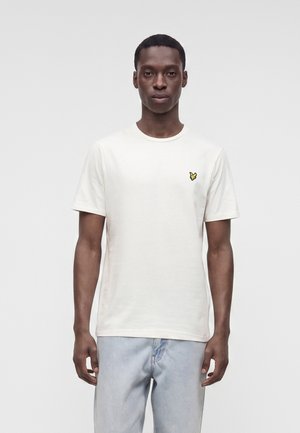 Homme portant un t-shirt blanc uni à manches courtes avec un petit logo d'oiseau jaune sur la poitrine gauche et un jean bleu clair.
