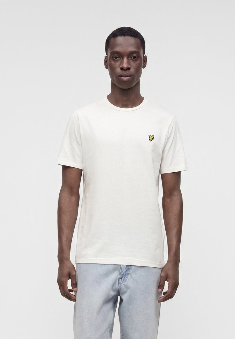 Homme portant un t-shirt blanc uni à manches courtes avec un petit logo d'oiseau jaune sur la poitrine gauche et un jean bleu clair.