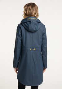Vattentät marinblå regnkappa med huva, har gula detaljer, justerbar midja och texten "WENN DAT FLADDERT" på kragen.