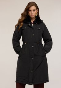 Svart lång parkasjacka med fuskpälskrage, dragsko i midjan, framfickor och tryckknappar. Slät textur och avslappnad passform.