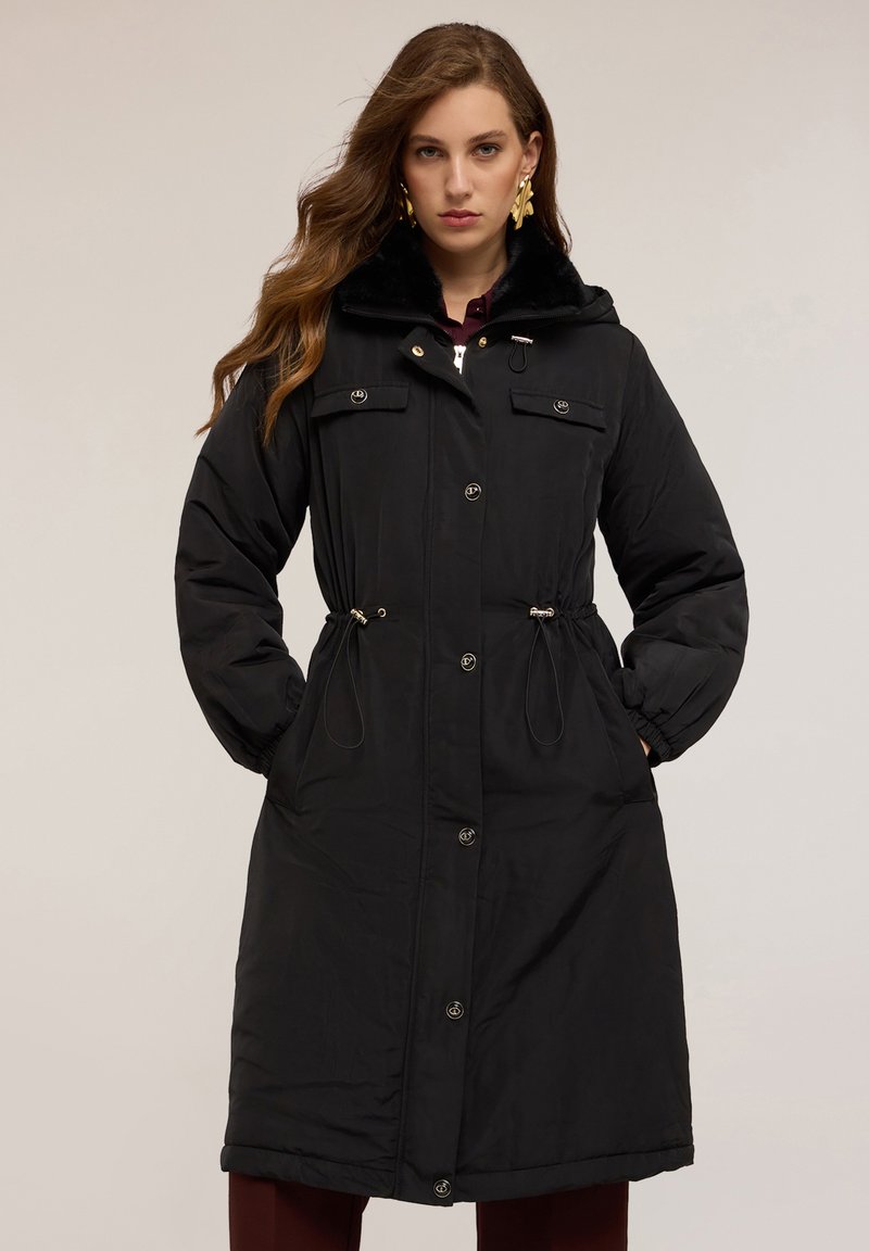 Svart lång parkasjacka med fuskpälskrage, dragsko i midjan, framfickor och tryckknappar. Slät textur och avslappnad passform.