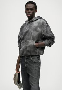 Μαύρο πουλόβερ tie-dye με τσέπη στο μπροστινό μέρος, φορεμένο με σκούρα τζιν. Ένα γκρι καπέλο κρατιέται στο χέρι; η υφή του υφάσματος δημιουργεί μια άνετη εμφάνιση.