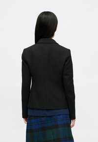 Črn fitted blazer z izrezom na lapelah, prilagojenim zadnjim šivom, nošen preko modre rebraste majice, v kombinaciji s karirasto krilo v zeleni in modri barvi.