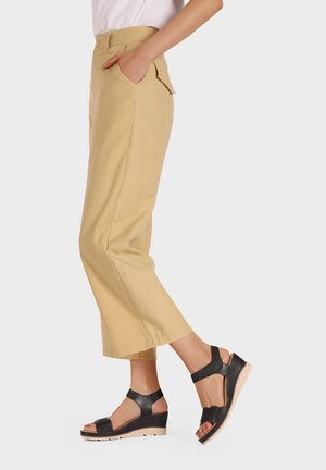 Pantaloni beige a vita alta, tagliati, con tasche laterali, abbinati a sandali neri con zeppa caratterizzati da cinturini texturizzati e una suola di colore chiaro.