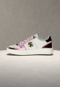 Λευκό αθλητικό παπούτσι με ροζ μεταλλικές λεπτομέρειες υφής φίδι και χρυσό λογότυπο στο πλάι, τοποθετημένο σε ανοιχτή επιφάνεια με ουδέτερο φόντο.