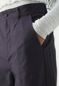 Jūros mėlynumo smulkiai dryžuoti kelnių su šoninėmis kišenėmis, užsegimu mygtuku ir tekstūruotu paviršiumi, derinami su balta ilgomis rankovėmis marškiniais.