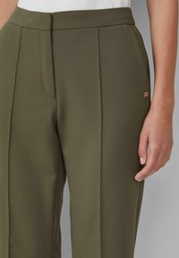 Pantaloni su misura verde oliva realizzati in tessuto liscio, con taglio dritto, pieghe frontali e un piccolo dettaglio dorato sulla tasca.