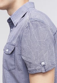 Gros plan d'une personne portant une chemise à manches courtes à carreaux bleus et blancs avec une poche poitrine boutonnée et un col.