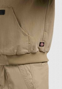Gros plan sur la poche d'une veste beige et son ourlet côtelé au-dessus d'un pantalon assorti, montrant l'étiquette de la marque Khujo sur la couture latérale de la veste.
