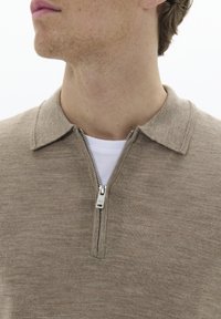 Camisa polo de color marrón claro con cremallera, de punto texturizado, que presenta un cuello tipo solapa y un detalle de cremallera plateada, superpuesta sobre una camisa blanca.