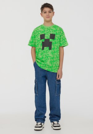 Maglietta verde pixelata a maniche corte con design di un volto di creeper di Minecraft nero, abbinata a pantaloni cargo blu e sneakers nere.