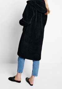 Czarny, puszysty płaszcz z kapturem o krótkich rękawach, długości do kolan, noszony na niebieskie dżinsy i czarne wkładane buty. Miękka faktura i prosty styl.
