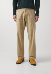 Man som bär beige byxor, senapsgula mockaskor och en mörkgrön långärmad skjorta stående framför en enfärgad vit bakgrund.