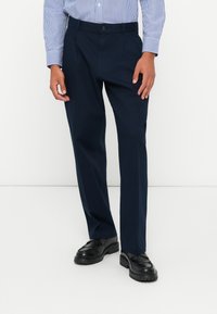 Jūras spēku bikses, izgatavotas no gludas auduma, ar taisnu kāju dizainu, priekšējām kabatām un pogāmas aizdare, apvienotas ar melnām kurpēm.