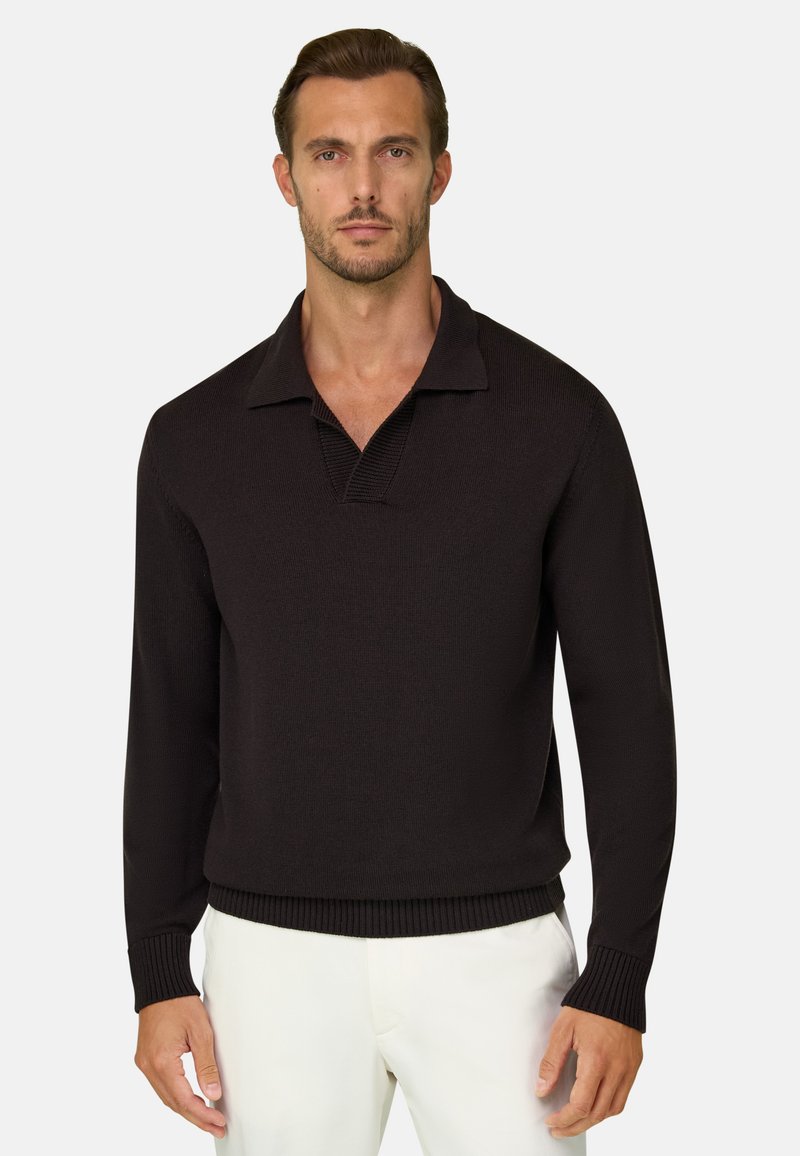 Homme portant un pull en maille noir à manches longues avec col et pantalon blanc, debout devant un fond clair uni.