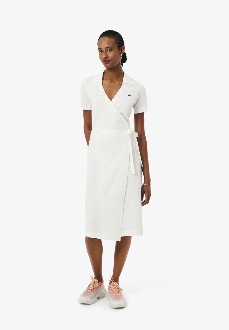 Femme debout portant une robe portefeuille blanche à manches courtes et col, assortie à des baskets épaisses roses et blanches.