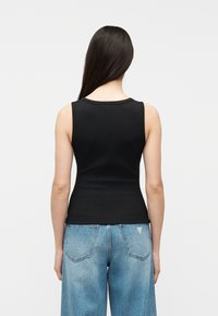 Femme aux longs cheveux noirs portant un top noir sans manches ajusté et un jean bleu ample, debout de dos devant un fond uni.