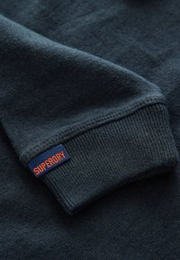 Mörkblå tröjdetalj med ribbad mudd och ett tygmärke med texten "SUPERDRY" i orange. Mjuk textur, avslappnad design.