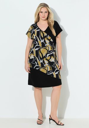 Kvinde iført knælange sort kjole med guld-, hvid- og sort abstrakt bladmønster, V-udskæring og korte ærmer, kombineret med sorte hæle.