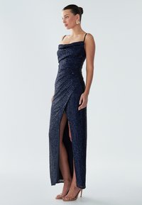 Navy sequined evening gown with a cowl neck, asymmetric draping, and a thigh-high slit. Styled with strappy nude high-heel sandals.