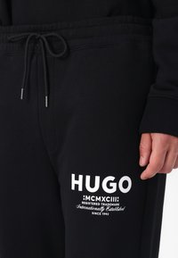 Black jogger pants made of cotton blend with an elastic waistband, drawstring closure, and white graphic logo on the lower leg.