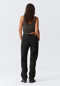 Pikkade lokkis pruunide juustega naine, kes kannab tumedat spagettite sifooni, musti sirgeid teksaseid ja musti kontsakingi, seismas valgel taustal.