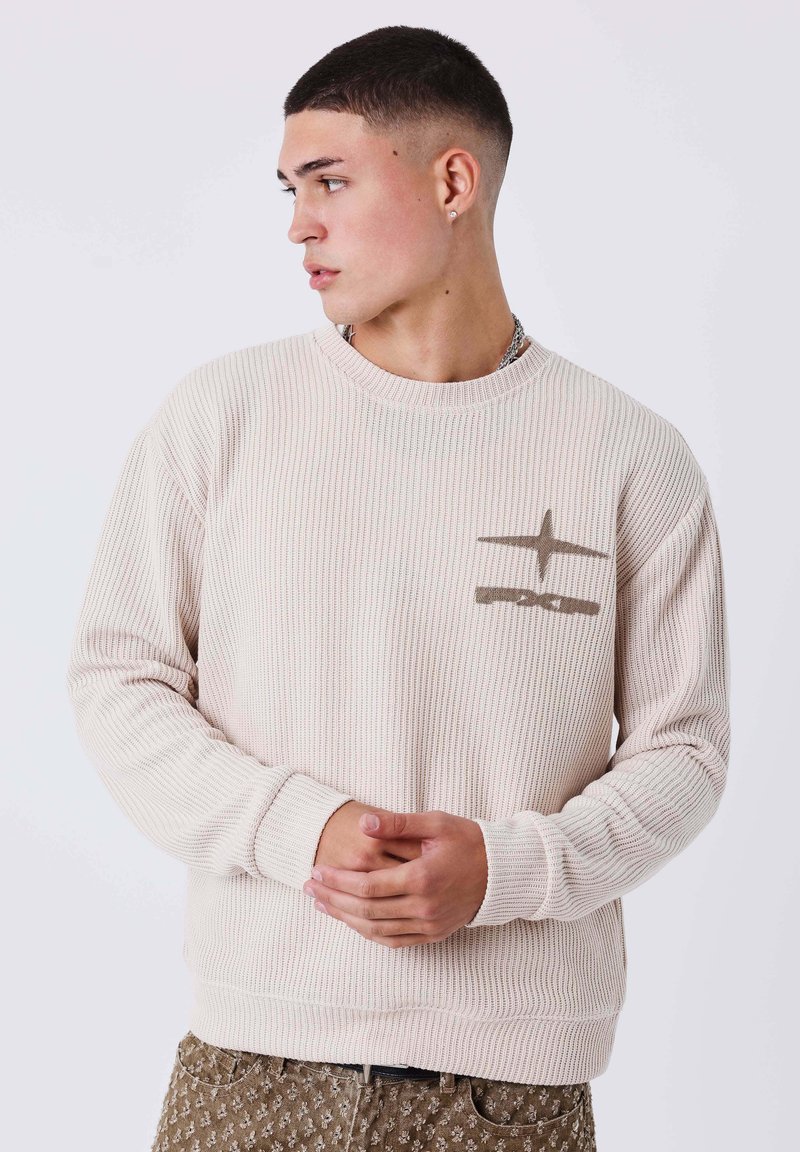 Joven con el cabello corto mira hacia la izquierda, viste un suéter de canalé beige con un diseño de estrella y pantalones marrones estampados, con las manos entrelazadas.