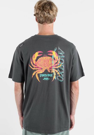 Personne portant un t-shirt gris foncé avec un grand crabe rouge et jaune ainsi que les mots « STOKED SINCE 1985 » et « OXYBOW » au dos.