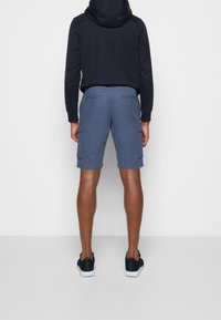 Blaue Cargo-Shorts mit Seitentaschen, getragen mit einem schwarzen Hoodie. Sie haben eine glatte Textur und einen geraden Schnitt, kombiniert mit schwarzen Sneakers.