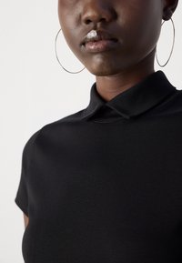Zwarte polo met een kraag, korte mouwen en een gladde textuur. Zilveren ringoorbellen zichtbaar langs de halslijn.